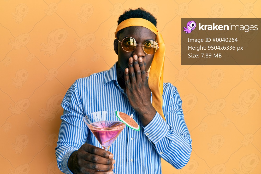 Handsome black man drunk wearing tie over head and sunglasses drinking a cocktail covering mouth with hand, shocked and afraid for mistake. surprised expression 
