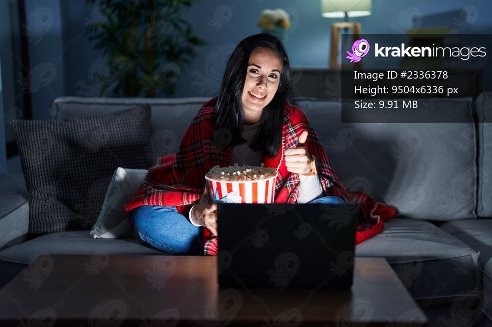 Hispanic woman eating popcorn watching a movie on the sofa doing happy thumbs up gesture with hand. approving expression looking at the camera showing success. 