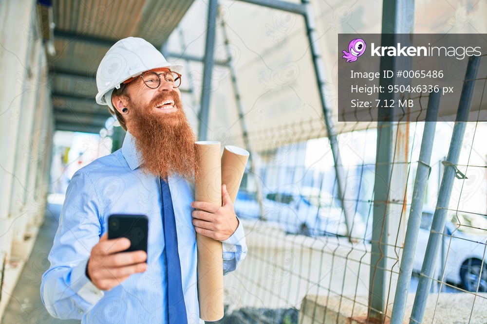 Young redhead architect man with long beard wearing hardhat smiling happy. Holding blueprints using smartphone at street of city.