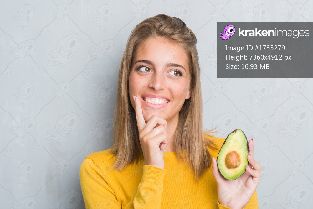 Beautiful young woman over grunge grey wall eating avocado serious face thinking about question, very confused idea
