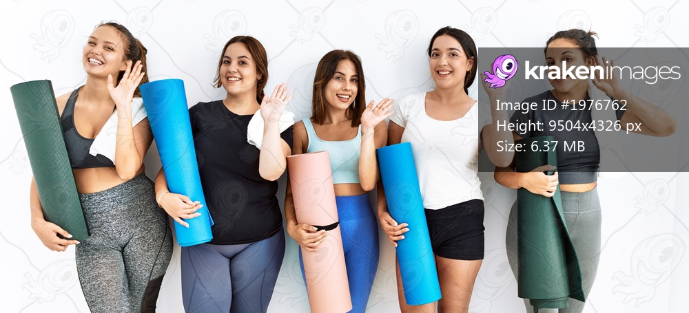 Group of women holding yoga mat standing over isolated background waiving saying hello happy and smiling, friendly welcome gesture 