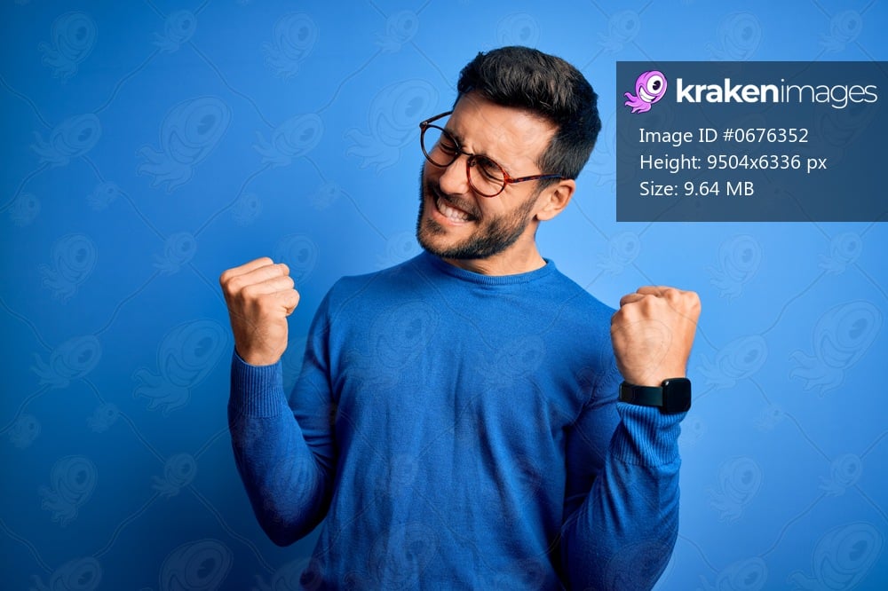 Young handsome man with beard wearing casual sweater and glasses over blue background very happy and excited doing winner gesture with arms raised, smiling and screaming for success. Celebration concept.