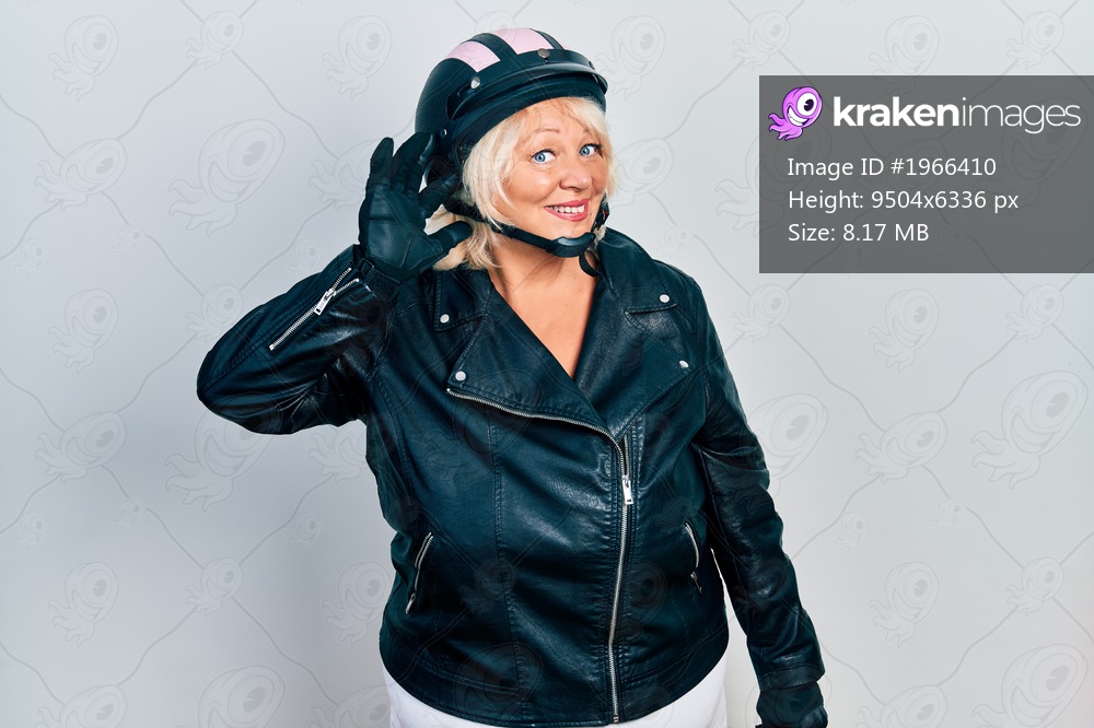 Middle age blonde woman holding motorcycle helmet smiling with hand over ear listening an hearing to rumor or gossip. deafness concept. 