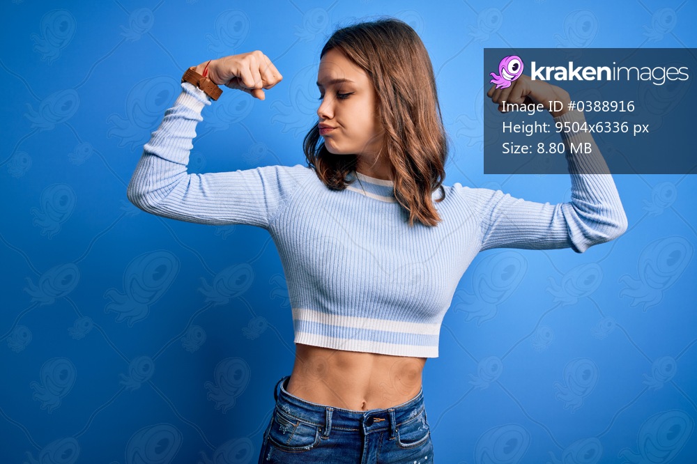 Young beautiful blonde girl wearing casual sweater standing over blue isolated background showing arms muscles smiling proud. Fitness concept.