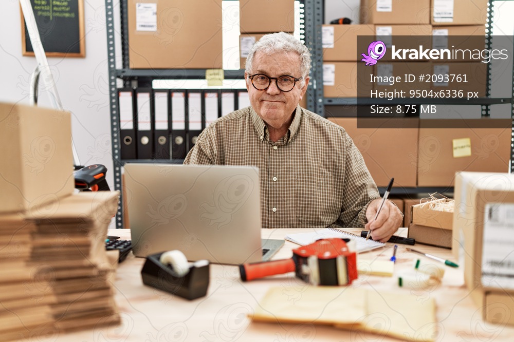 Middle age grey-haired man using laptop and writing on notebook working at storehouse