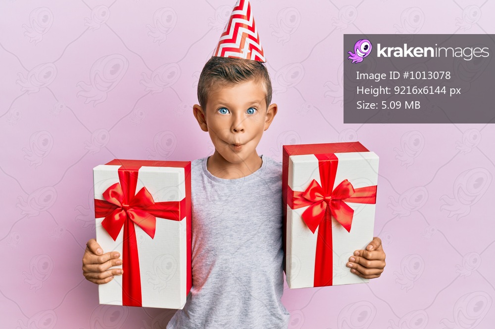 Adorable caucasian kid wearing birthday hat holding presents making fish face with mouth and squinting eyes, crazy and comical. 
