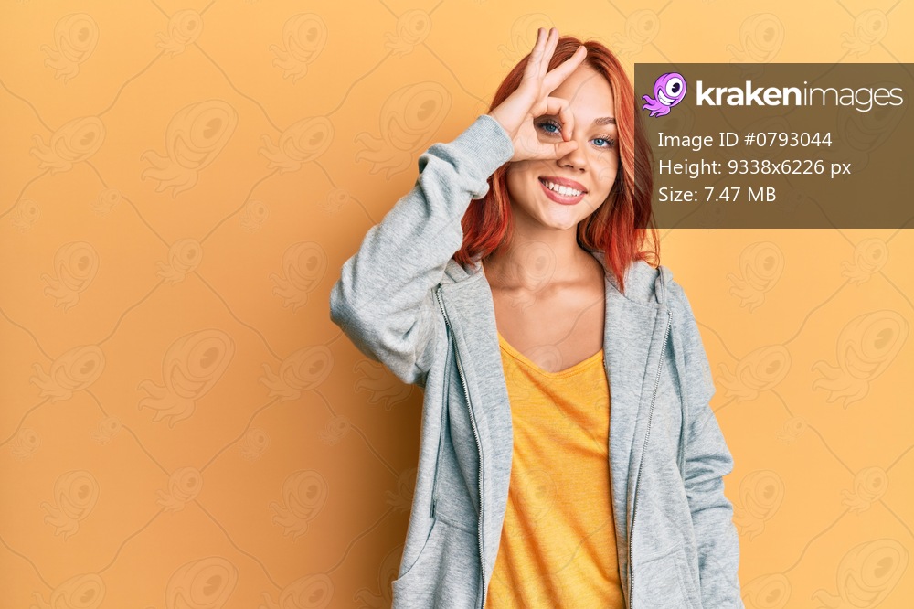 Young beautiful redhead woman wearing casual sporty sweatshirt smiling happy doing ok sign with hand on eye looking through fingers 