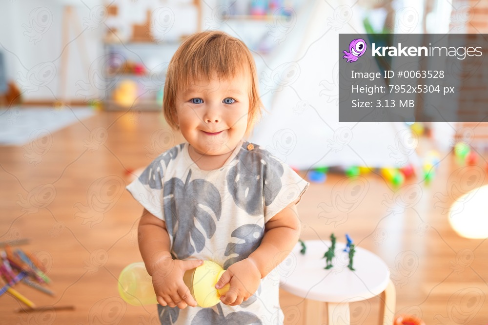 Adorable toddler standing with smile on face holding feeding bottle around lots of toys at kindergarten