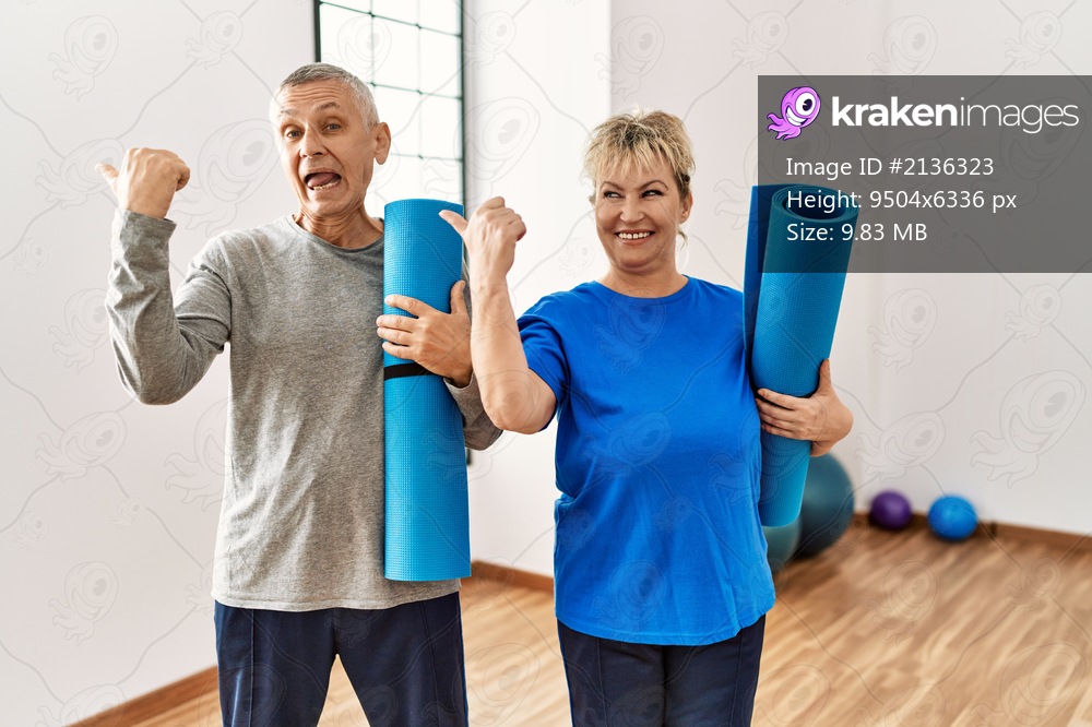 Middle age caucasian couple holding yoga mat at pilates room pointing thumb up to the side smiling happy with open mouth 