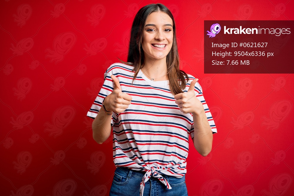 Young beautiful brunette woman wearing casual striped t-shirt standing over red background success sign doing positive gesture with hand, thumbs up smiling and happy. Cheerful expression and winner gesture.