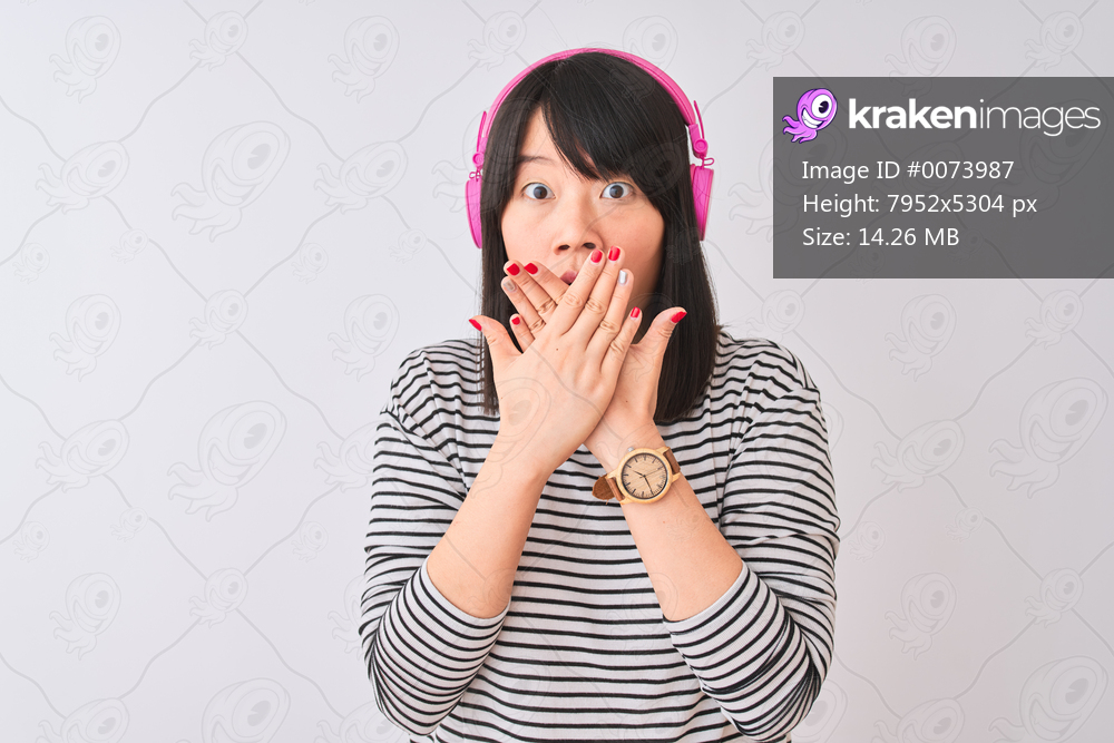 Chinese woman listening to music using pink headphones over isolated white background shocked covering mouth with hands for mistake. Secret concept.