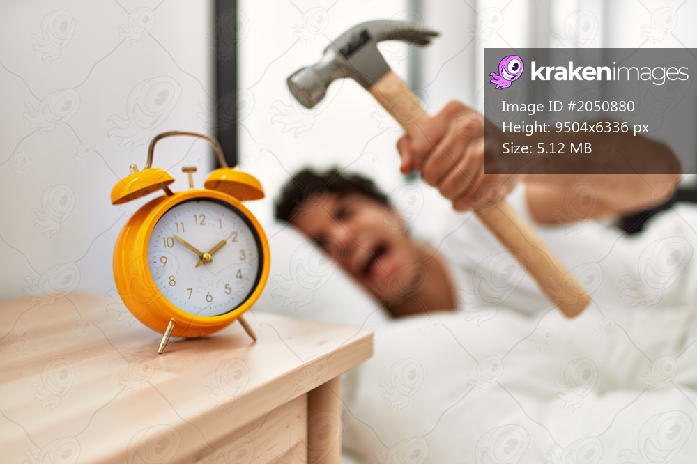 Young hispanic man turning off alarm clock using hammer lying on the bed at bedroom.