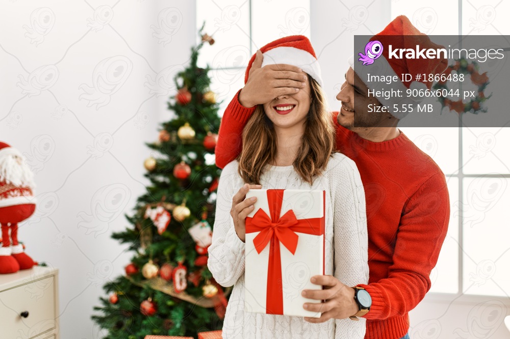 Young hispanic couple wearing christmas hat. Man surprising his girlfriend with hands on eyes and gift at home.