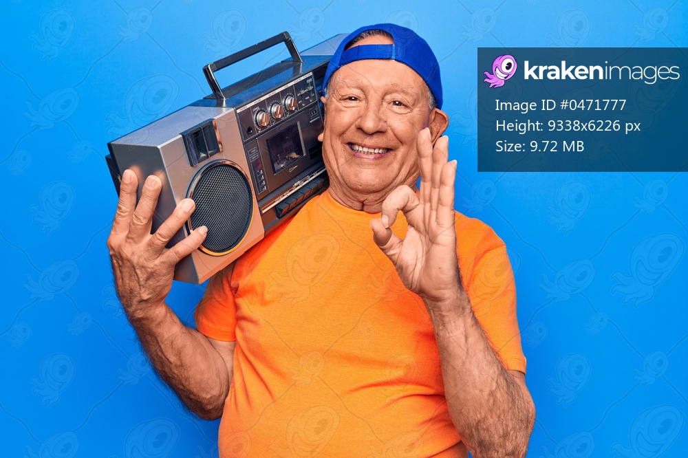 Senior modern handsome grey-haired man wearing cap listening to music holding boombox doing ok sign with fingers, smiling friendly gesturing excellent symbol