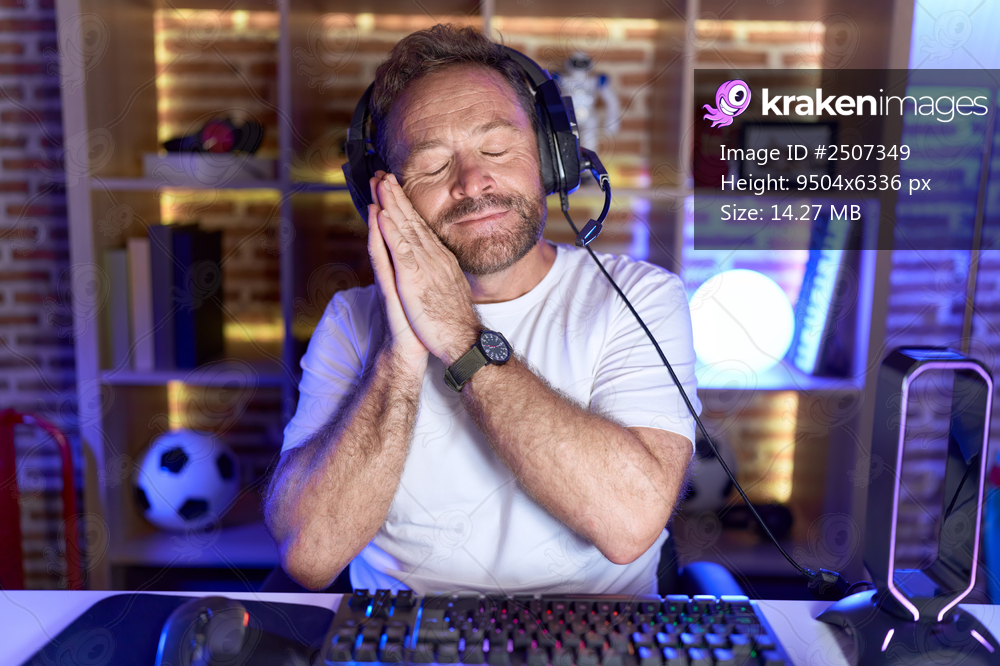 Middle age man with beard playing video games wearing headphones sleeping tired dreaming and posing with hands together while smiling with closed eyes. 