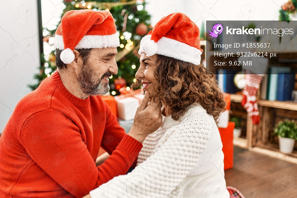 Middle age hispanic couple wearing christmas hat sitting on the floor and hugging at home.