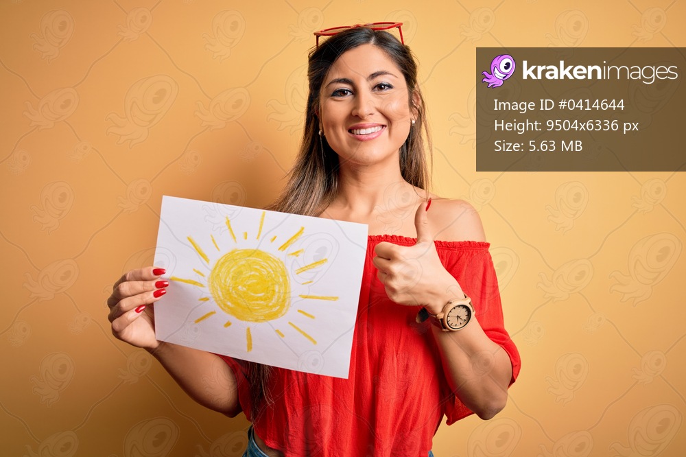 Young beautiful brunette woman holding paper with sun draw over isolated yellow background happy with big smile doing ok sign, thumb up with fingers, excellent sign