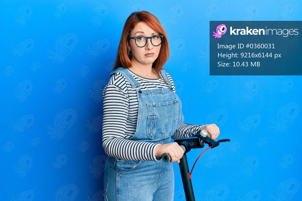 Beautiful redhead woman standing on modern scooter over blue background skeptic and nervous, frowning upset because of problem. negative person. 