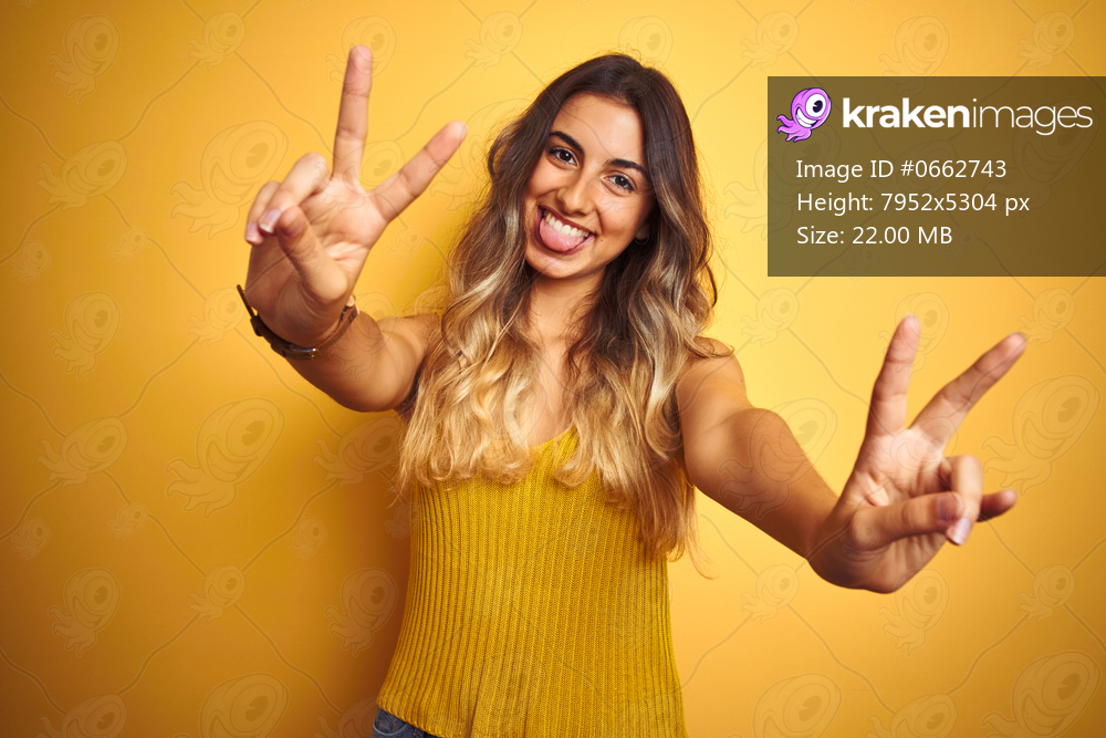 Young beautiful woman wearing t-shirt over yellow isolated background smiling with tongue out showing fingers of both hands doing victory sign. Number two.