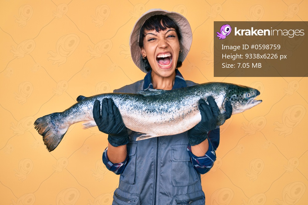 Beautiful brunettte fisher woman showing raw salmon celebrating crazy and amazed for success with open eyes screaming excited. 