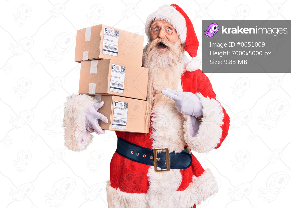 Old senior man with grey hair and long beard wearing santa claus costume holding boxes smiling happy pointing with hand and finger 