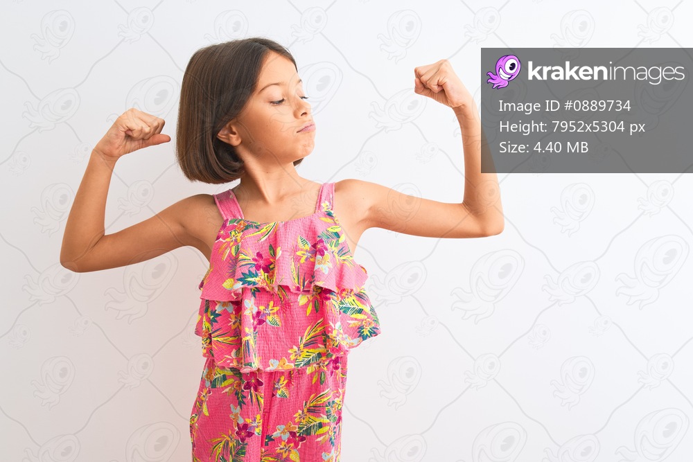 Young beautiful child girl wearing pink floral dress standing over isolated white background showing arms muscles smiling proud. Fitness concept.