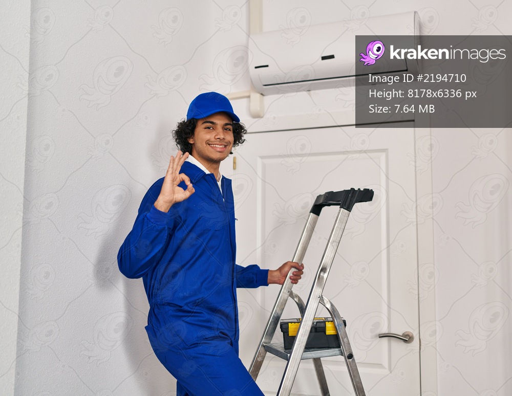 Hispanic man with curly hair working at home renovation doing ok sign with fingers, smiling friendly gesturing excellent symbol 