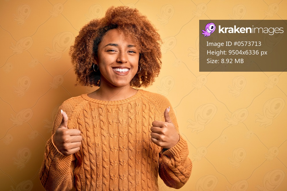 Young beautiful African American afro woman with curly hair wearing casual sweater success sign doing positive gesture with hand, thumbs up smiling and happy. Cheerful expression and winner gesture.