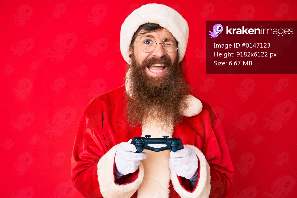 Handsome young red head man with long beard wearing santa claus costume playing video games winking looking at the camera with sexy expression, cheerful and happy face. 