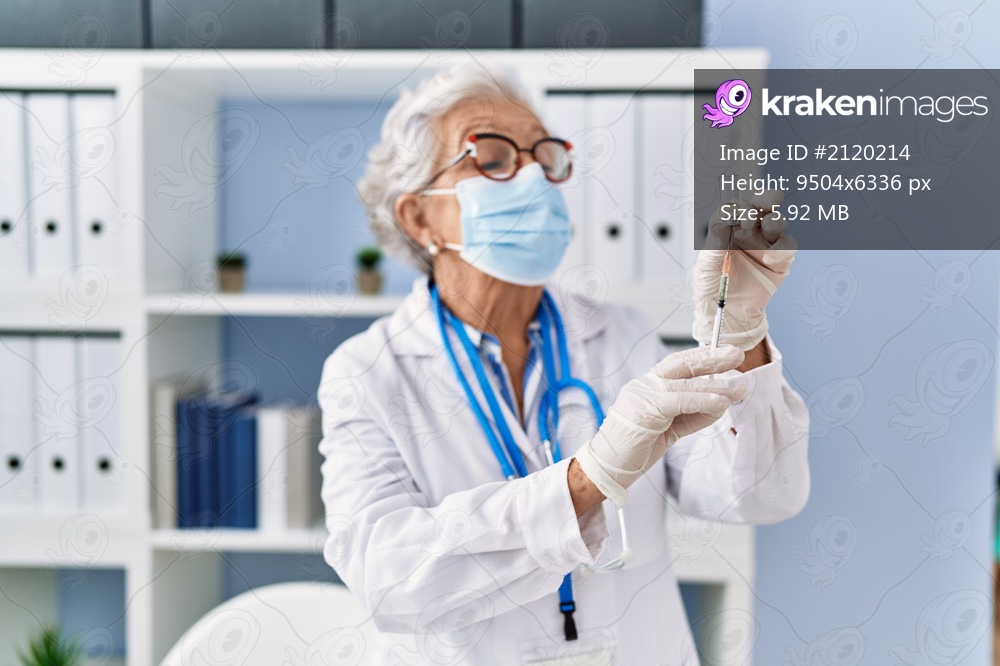 Senior grey-haired woman wearing doctor uniform and medical mask holding vaccine dose at clinic