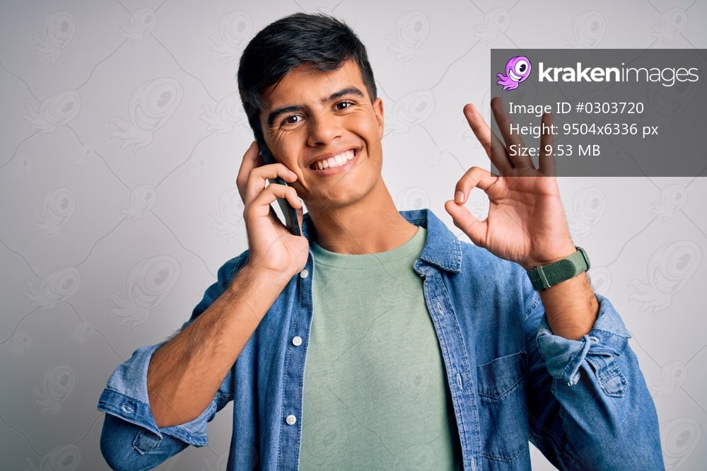 Young handsome man having conversation talking on the smartphone over white background doing ok sign with fingers, excellent symbol