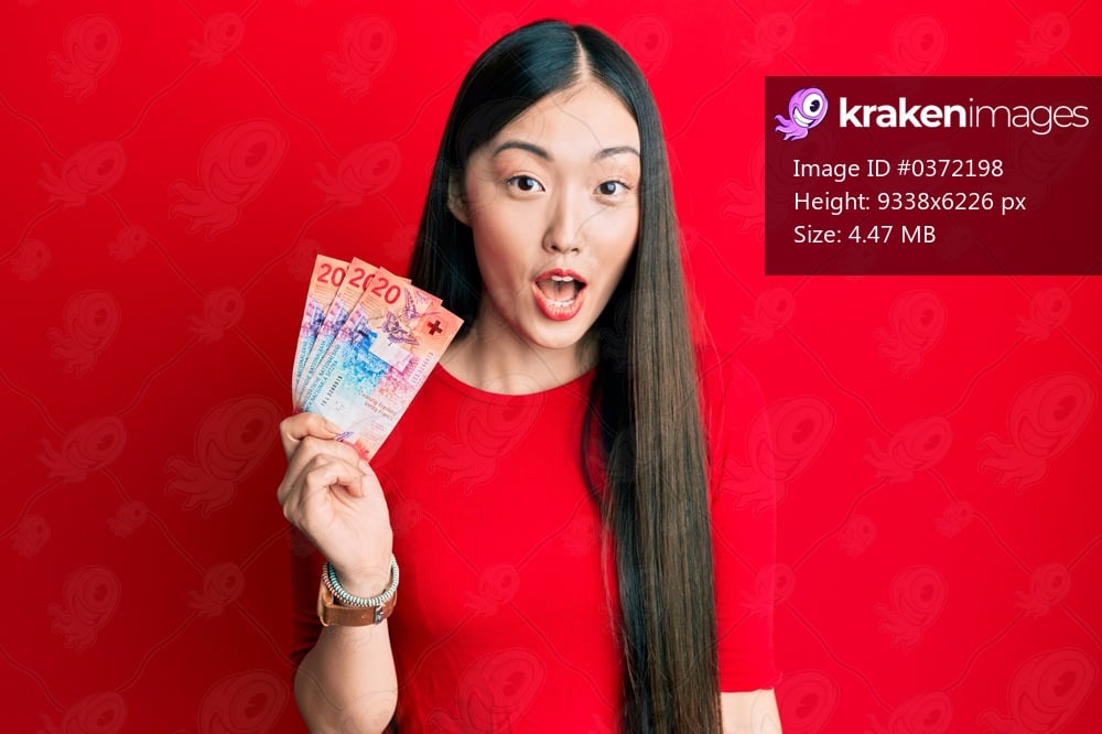 Young chinese woman holding 20 swiss franc banknotes scared and amazed with open mouth for surprise, disbelief face 