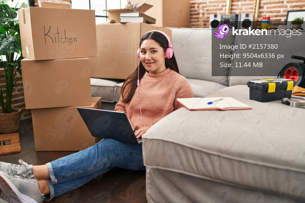 Young hispanic woman using laptop and headphones sitting on floor at new home