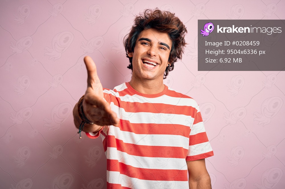 Young handsome man wearing striped casual t-shirt standing over isolated pink background smiling friendly offering handshake as greeting and welcoming. Successful business.