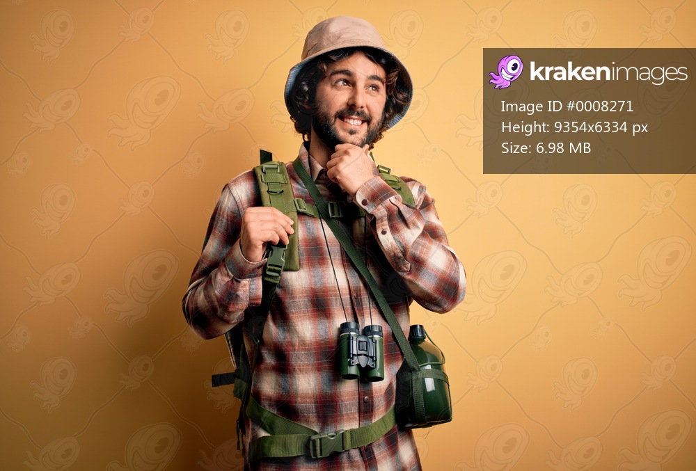 Young hiker man with curly hair and beard hiking wearing backpack and water canteen with hand on chin thinking about question, pensive expression. Smiling and thoughtful face. Doubt concept.