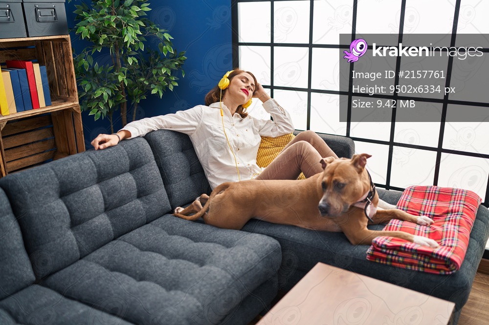 Young caucasian woman listening to music sitting on sofa with dog at home