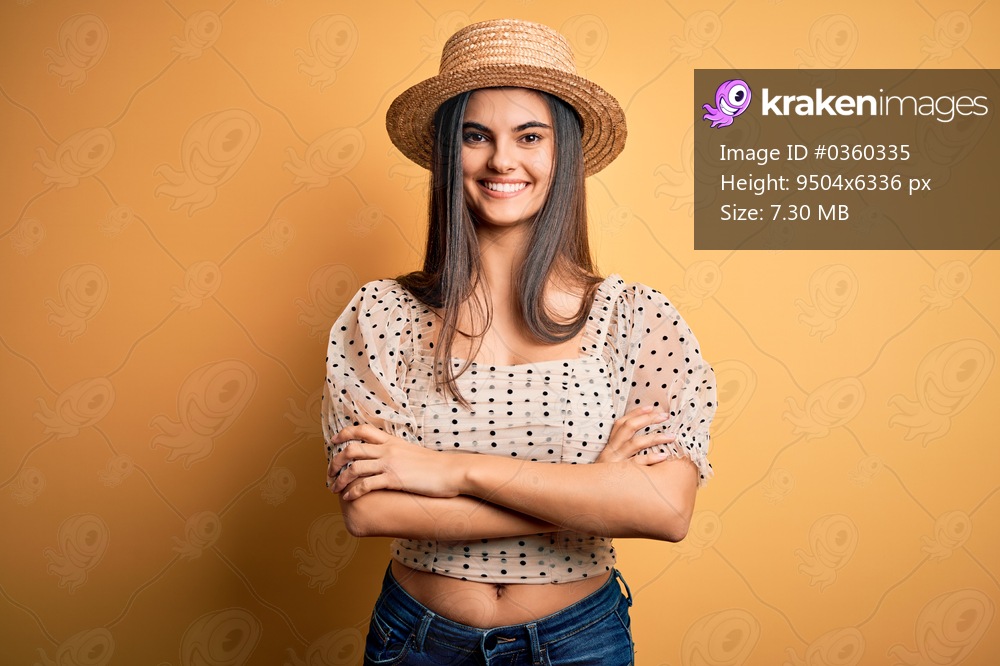 Young beautiful brunette woman wearing t-shirt and summer hat over yellow background happy face smiling with crossed arms looking at the camera. Positive person.