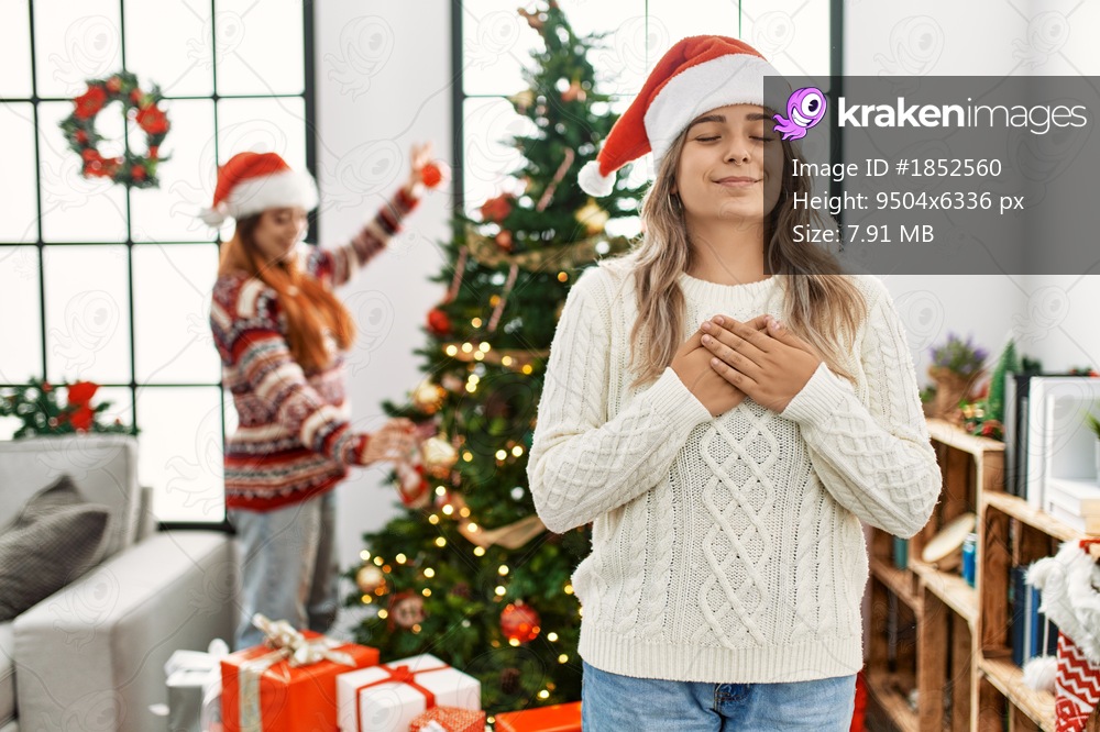 Beautiful couple at home standing by the christmas tree smiling with hands on chest with closed eyes and grateful gesture on face. health concept. 