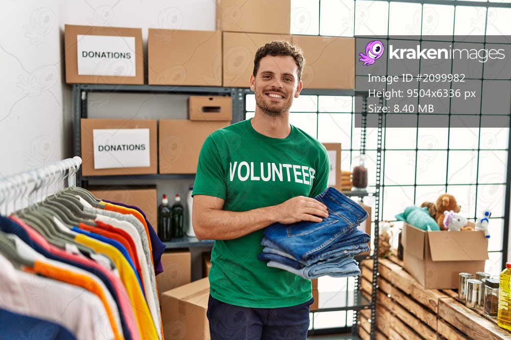 Young hispanic man wearing volunteer uniform holding folded jeans at charity center