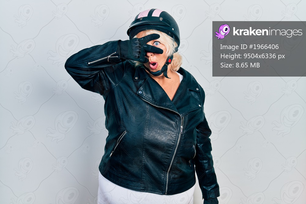 Middle age blonde woman holding motorcycle helmet peeking in shock covering face and eyes with hand, looking through fingers with embarrassed expression. 
