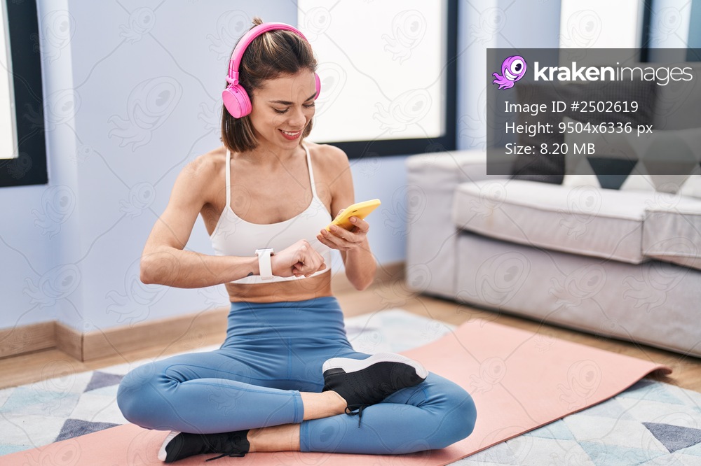 Young beautiful hispanic woman sitting on yoga mat listening to music looking stopwatch at home