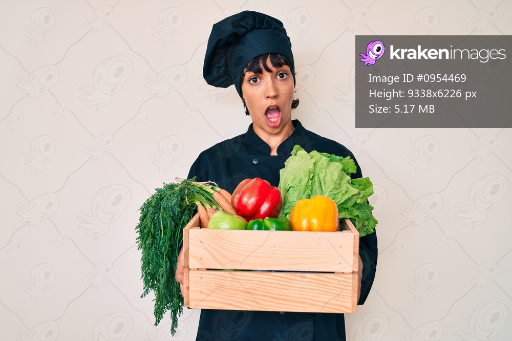 Beautiful brunettte woman chef holding fresh veggetables in shock face, looking skeptical and sarcastic, surprised with open mouth 