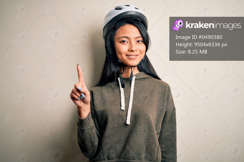 Young beautiful chinese woman wearing bike helmet over isolated white background showing and pointing up with finger number one while smiling confident and happy.