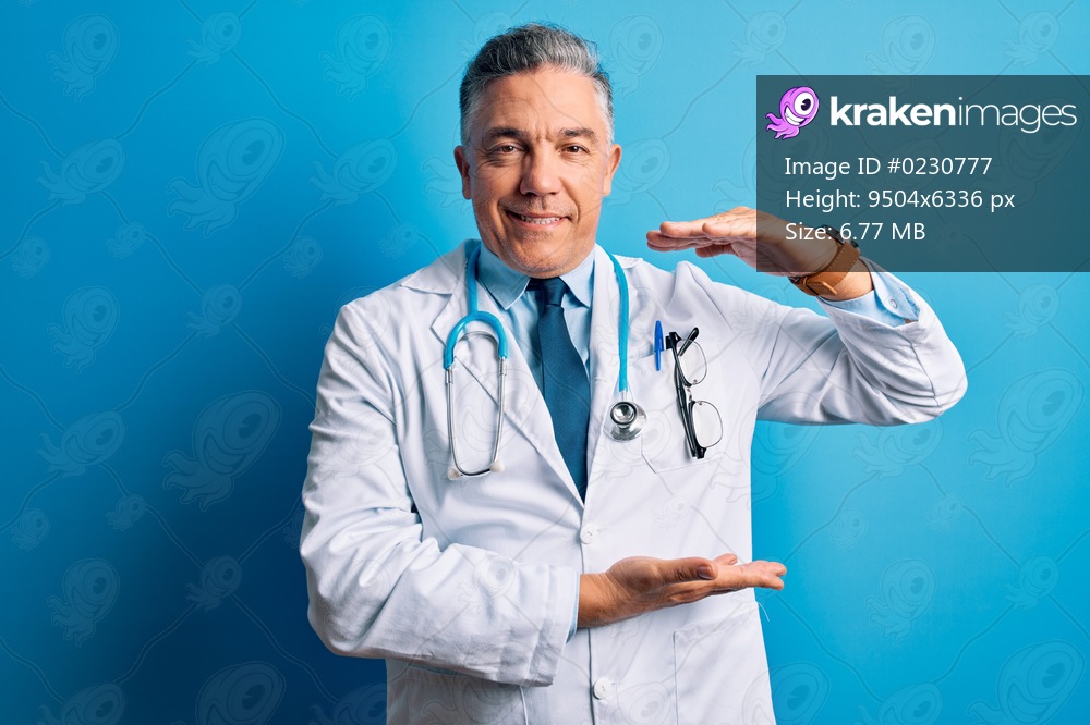 Middle age handsome grey-haired doctor man wearing coat and blue stethoscope gesturing with hands showing big and large size sign, measure symbol. Smiling looking at the camera. Measuring concept.