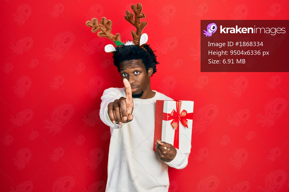 Young african american man wearing deer christmas hat holding gift pointing with finger up and angry expression, showing no gesture 