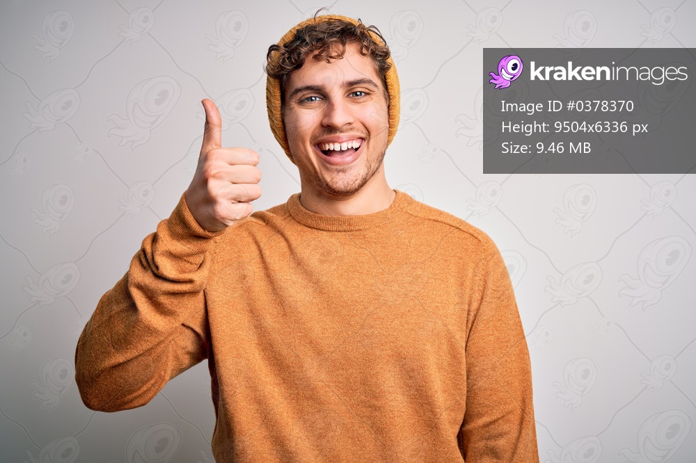 Young blond handsome man with curly hair wearing casual sweater and wool hat doing happy thumbs up gesture with hand. Approving expression looking at the camera showing success.