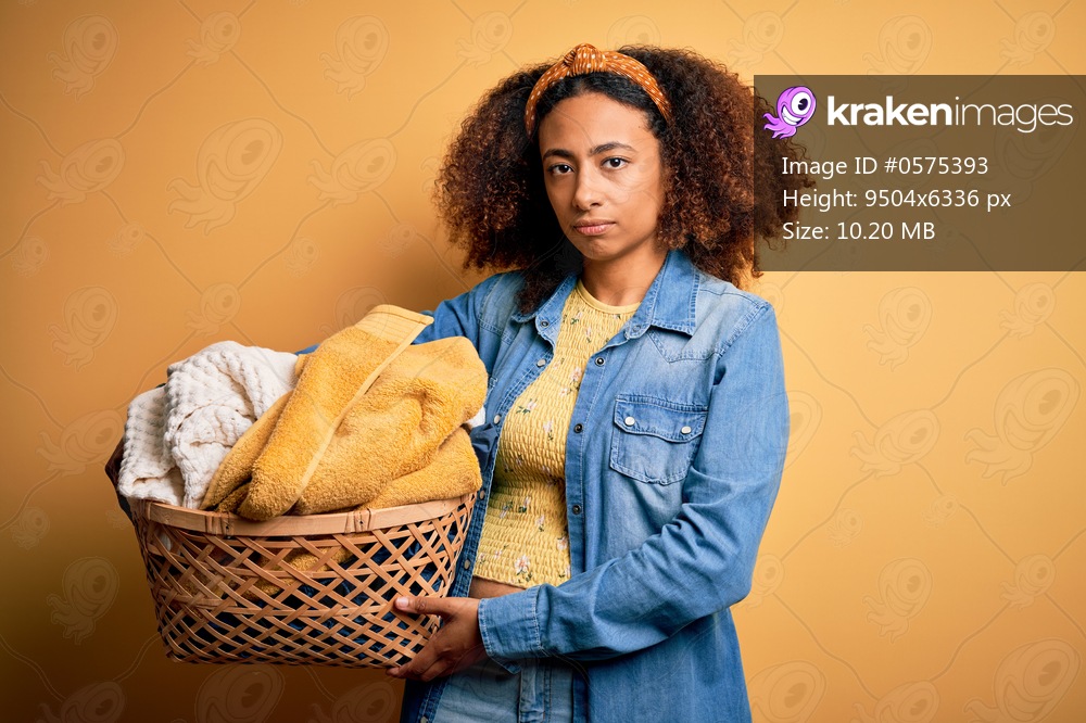 Young african american woman with afro hair holding wicker basket with clothes with a confident expression on smart face thinking serious