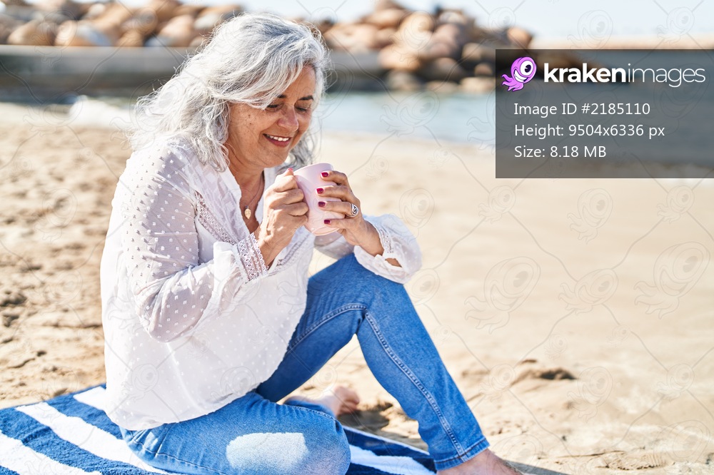 Middle age woman drinking coffee sitting on towel at seaside