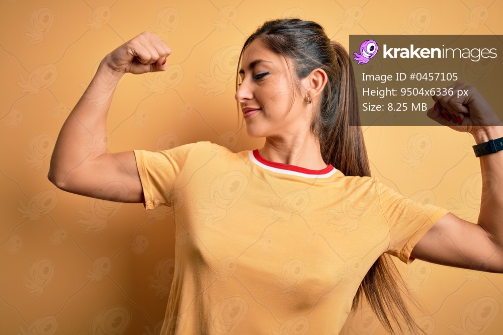 Young beautiful athletic woman wearing casual t-shirt and ponytail over yellow background showing arms muscles smiling proud. Fitness concept.