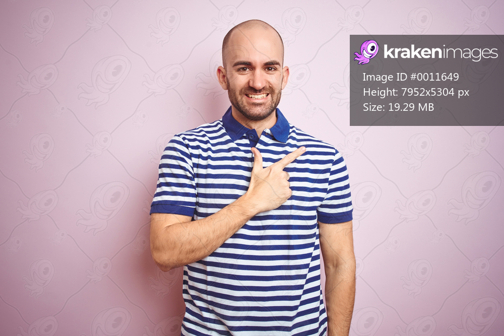Young bald man with beard wearing casual striped blue t-shirt over pink isolated background cheerful with a smile of face pointing with hand and finger up to the side with happy and natural expression on face